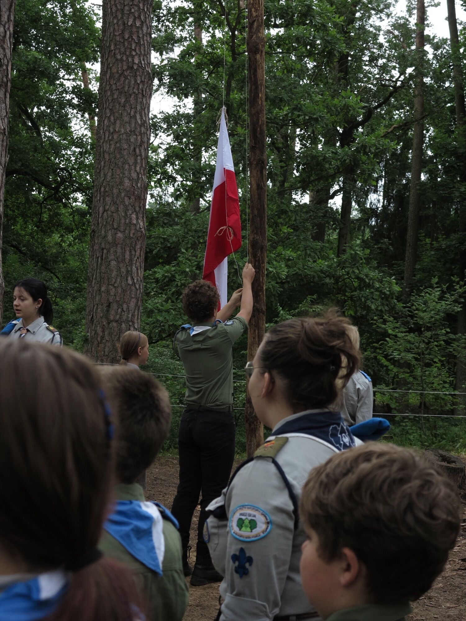 Harcerze w mundurach podnoszący biało-czerwoną flagę na maszt podczas obozu w lesie.