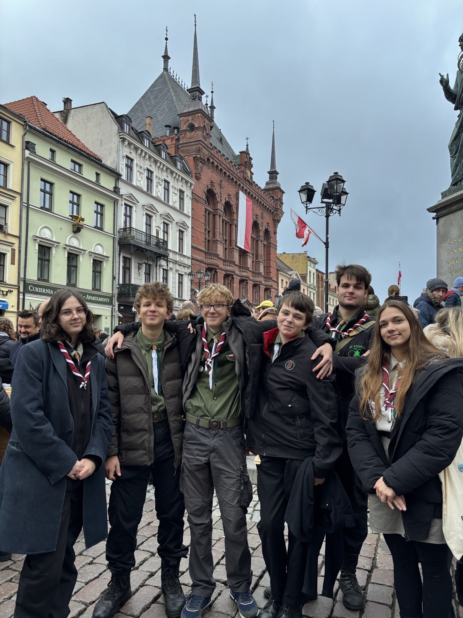 Grupa uśmiechniętych harcerzy w mundurach pozująca na tle zabytkowej architektury miasta.