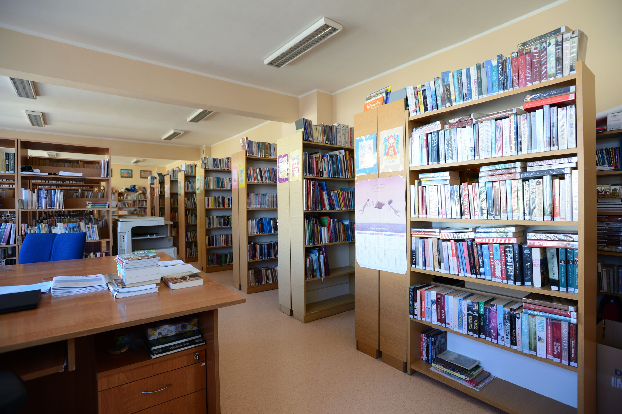 Wnętrze szkolnej biblioteki z jasnymi, drewnianymi regałami pełnymi książek oraz biurkiem.