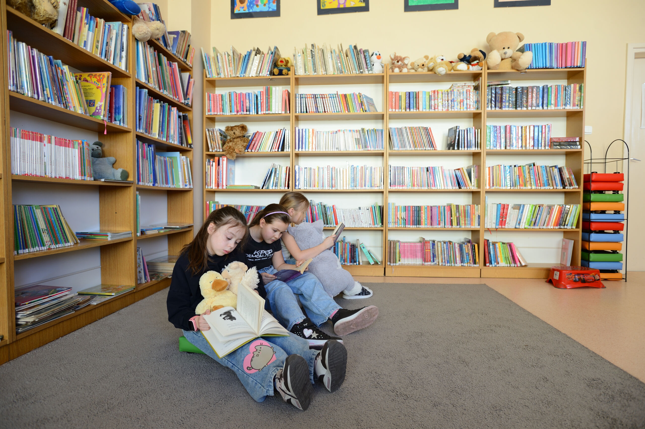 Trójka dzieci siedzących na dywanie w bibliotece, czytających książki w otoczeniu regałów z literaturą.