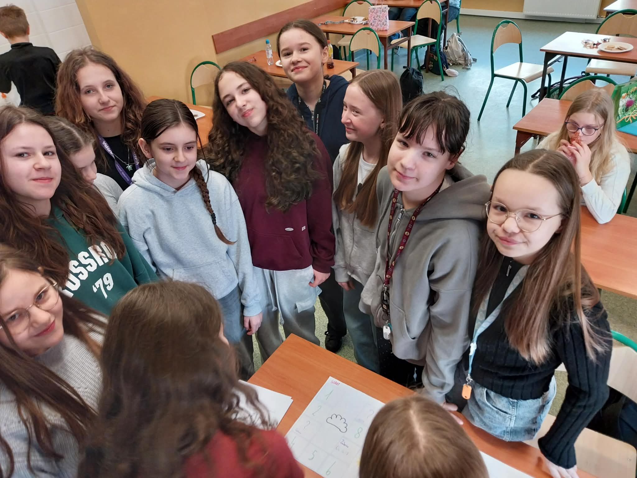 8ac7ce8d-4ba0-4499-92f7-0130253550c0 Grupa uśmiechniętych dziewczynek stojących wokół stolika z kartami i planszą w szkolnej klasie.