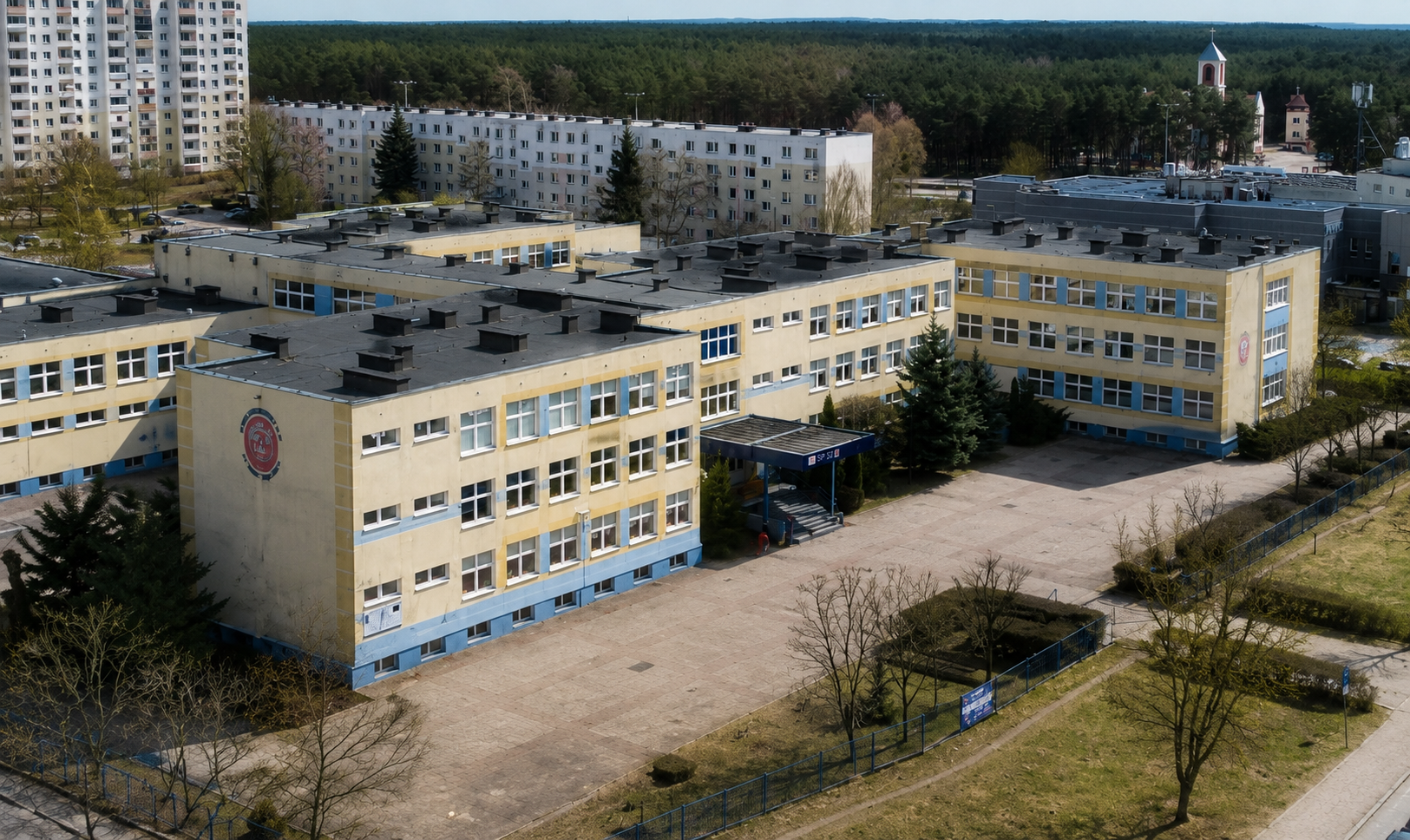 Widok z lotu ptaka na budynek Szkoły Podstawowej nr 32. Trzypiętrowy, jasnożółty gmach z niebieskimi elementami elewacji. Przed wejściem głównym znajduje się duży, utwardzony plac, a w tle widać pobliskie osiedle i las.