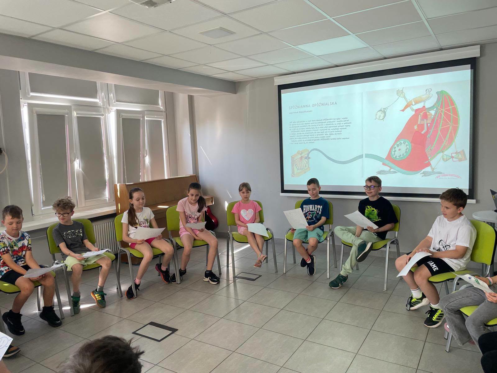 Grupa dzieci siedząca w półkolu na krzesłach w sali, patrząca na wyświetlaną z rzutnika stronę książki z ilustracją.