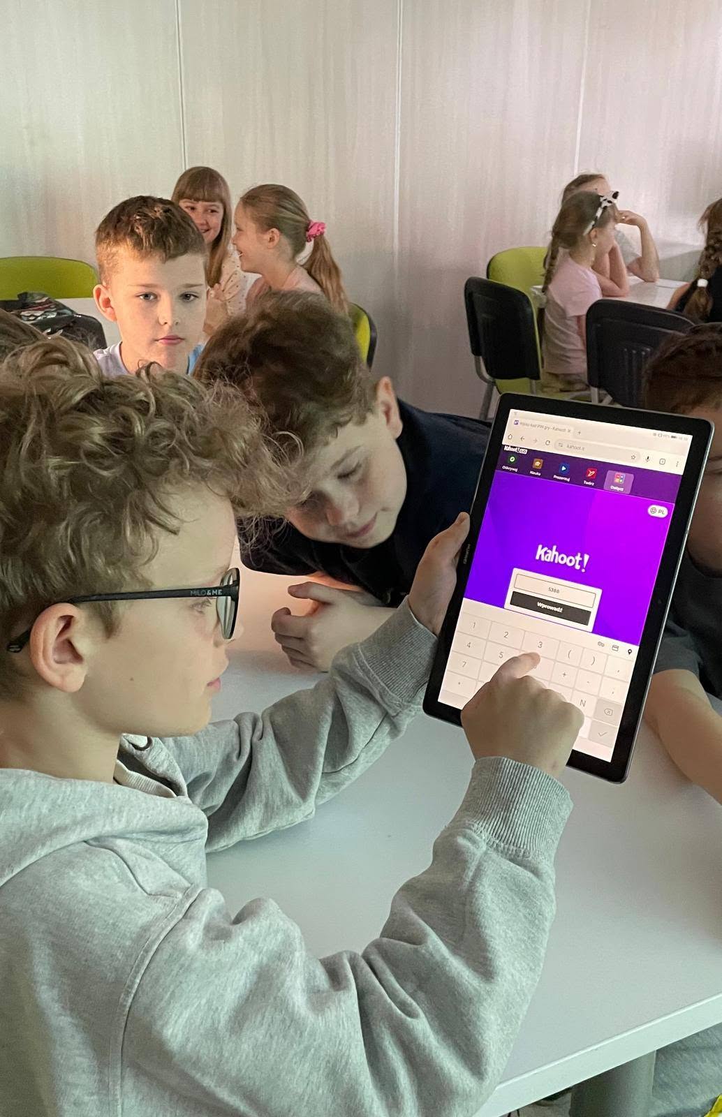 Chłopiec w okularach wpisujący kod do gry w aplikacji edukacyjnej Kahoot na tablecie, siedzący przy stoliku z rówieśnikami.