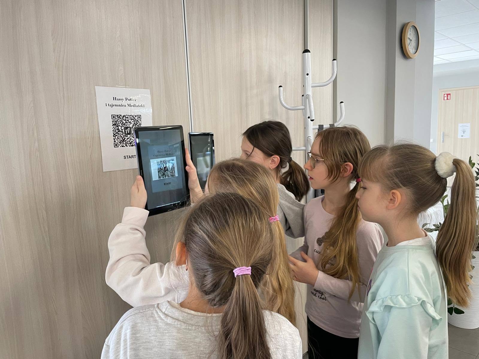 Grupa dziewczynek skanująca tabletem kod QR z kartki z napisem "Harry Potter i tajemnica Mediateki START".