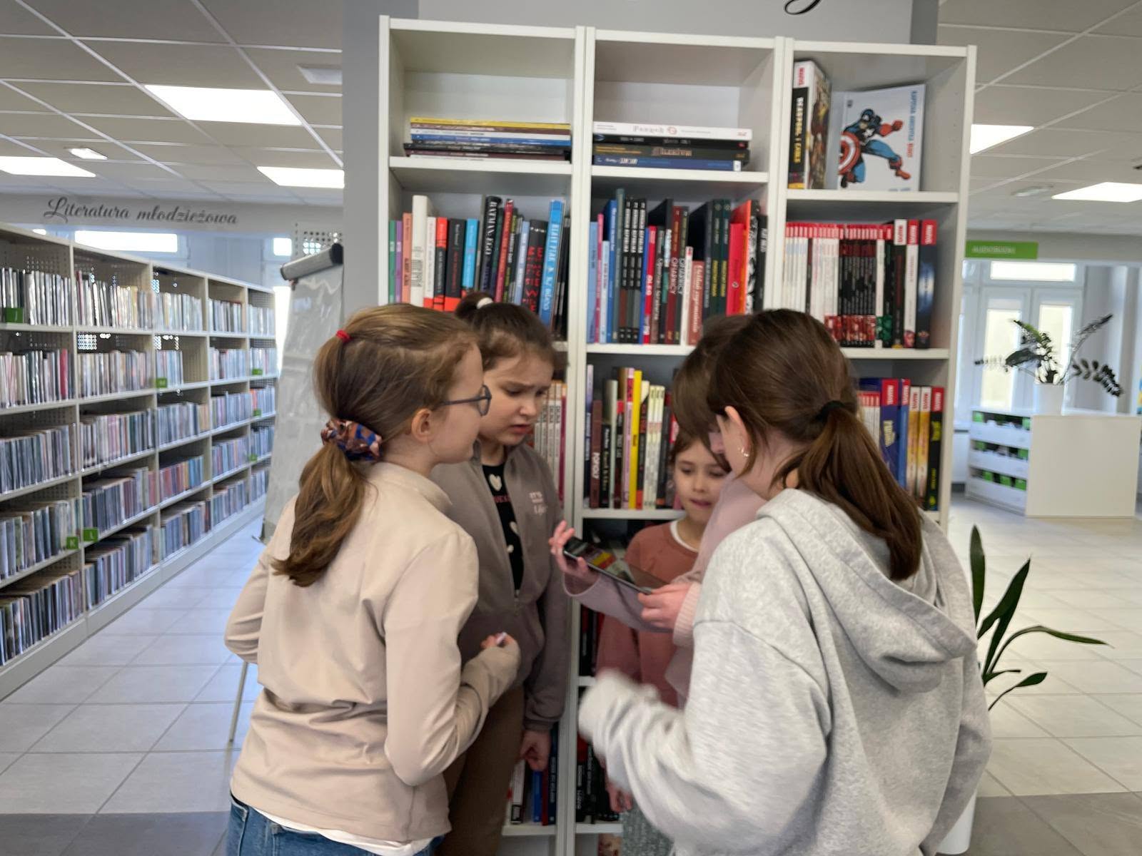 Cztery dziewczynki stojące w bibliotece przy regale z komiksami, wpatrzone w ekran tabletu.