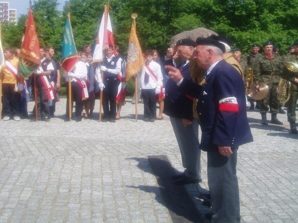 Kombatanci uczestniczący w uroczystości plenerowej w obecności szkolnych pocztów sztandarowych i orkiestry wojskowej. Jeden z mężczyzn salutuje.