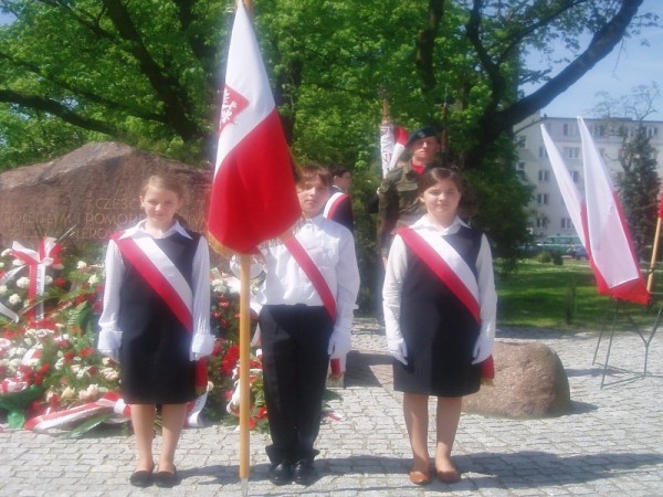Uczniowski poczet sztandarowy z polską flagą stojący przed kamiennym pomnikiem w asyście wojskowej.