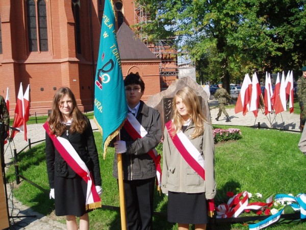 Uczniowski poczet sztandarowy trzymający sztandar szkoły podczas uroczystości na świeżym powietrzu. W tle rozwieszone polskie flagi.