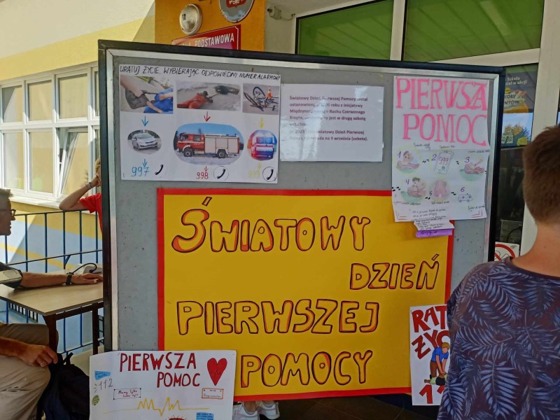 Tablica informacyjna z okazji Światowego Dnia Pierwszej Pomocy, zawierająca plakaty z instrukcjami ratunkowymi i numerami alarmowymi.