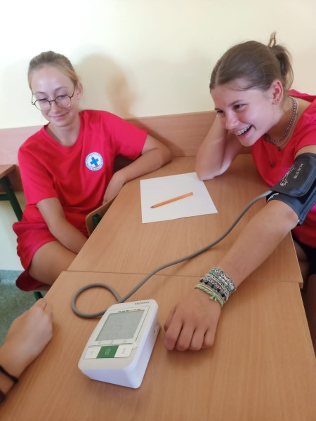 Dwie uśmiechnięte uczennice w ławce szkolnej podczas samodzielnego pomiaru ciśnienia krwi elektronicznym ciśnieniomierzem.