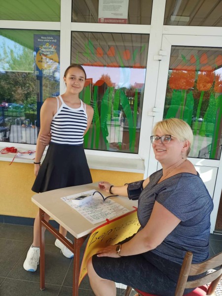 Uśmiechnięta kobieta mierząca ciśnienie uczennicy przy stoliku ustawionym przed wejściem do budynku.